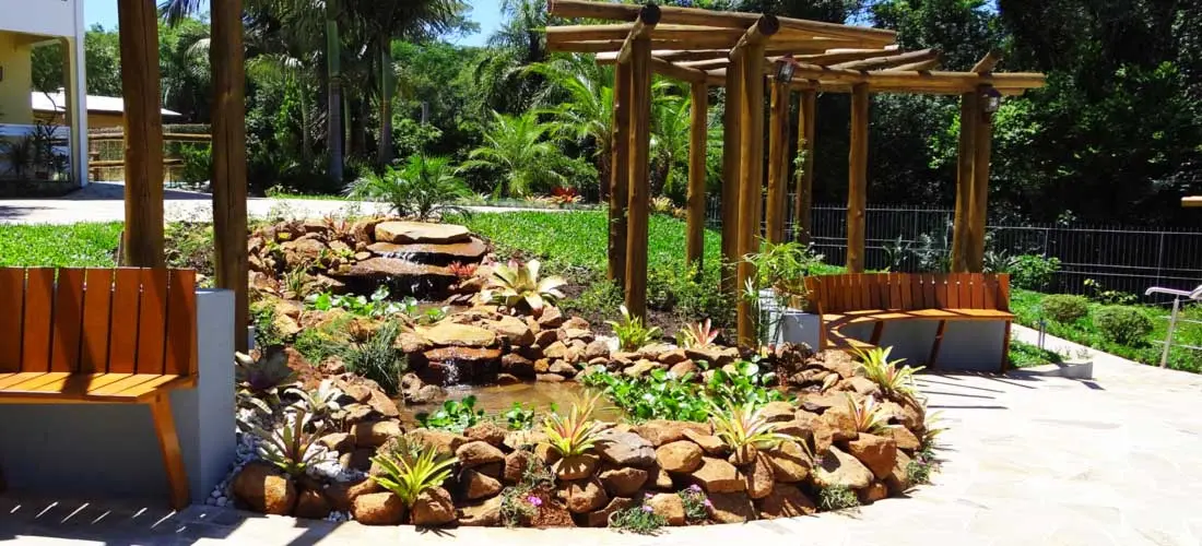 Jardim do Serraville com pergolado, bancos e fonte de pedras entre a vegetação