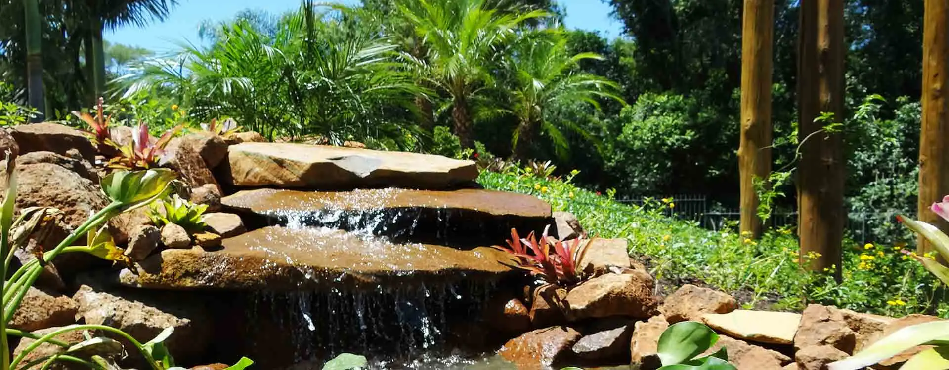 Fonte com cascata entre pedras, flores e palmeiras nos jardins do Serraville